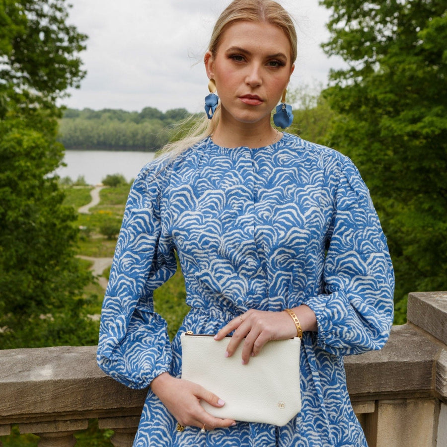 a stylish woman in a garden setting wearing a printed blue dress, carrying a white clutch purse with a chain wrist strap